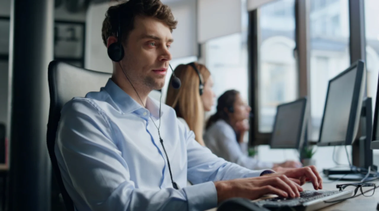 A man seen from the side, sitting at a desk with a headset on, typing on his PC.