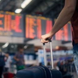 In the foreground, the arm and hand of a man holding a trolley; in the background, a departure board