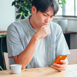Mann sitzt am Tisch mit einer Teetasse neben sich und schaut mit nachdenklichem Blick und einer Hand am Kinn auf sein Smartphone